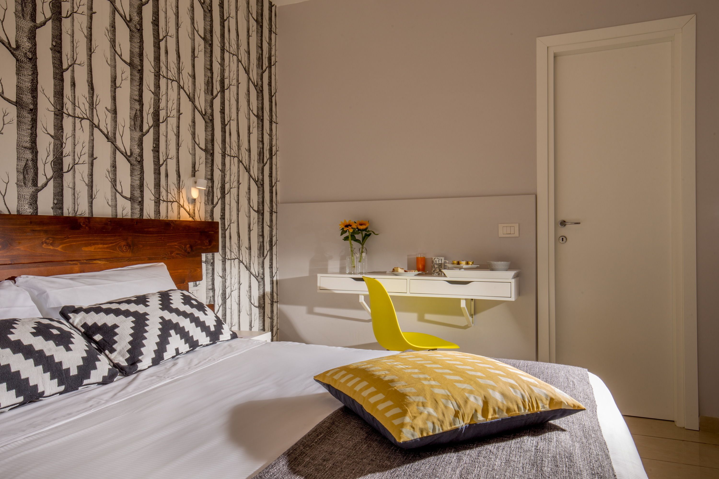 Quarto com cama, cabeceira de madeira, escrivaninha e cadeira, e uma porta.