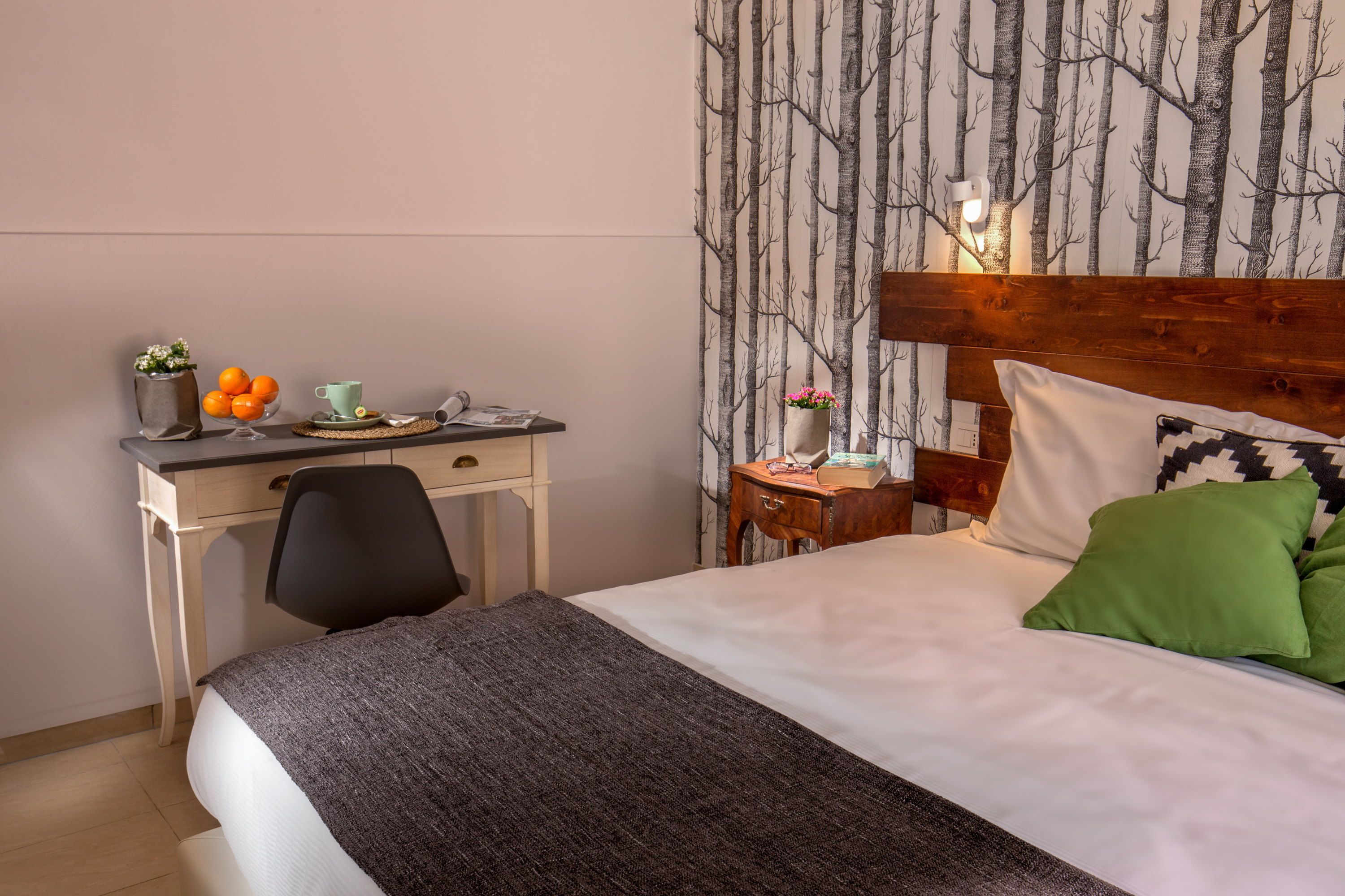 A hotel room with a bed, a desk, a chair, a nightstand, and a wall with a tree pattern.