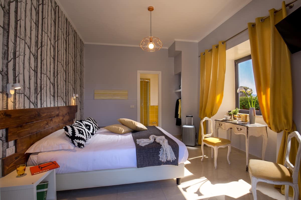 A bedroom with a bed, bedside table, lamp, and a view of the outdoors through a window.