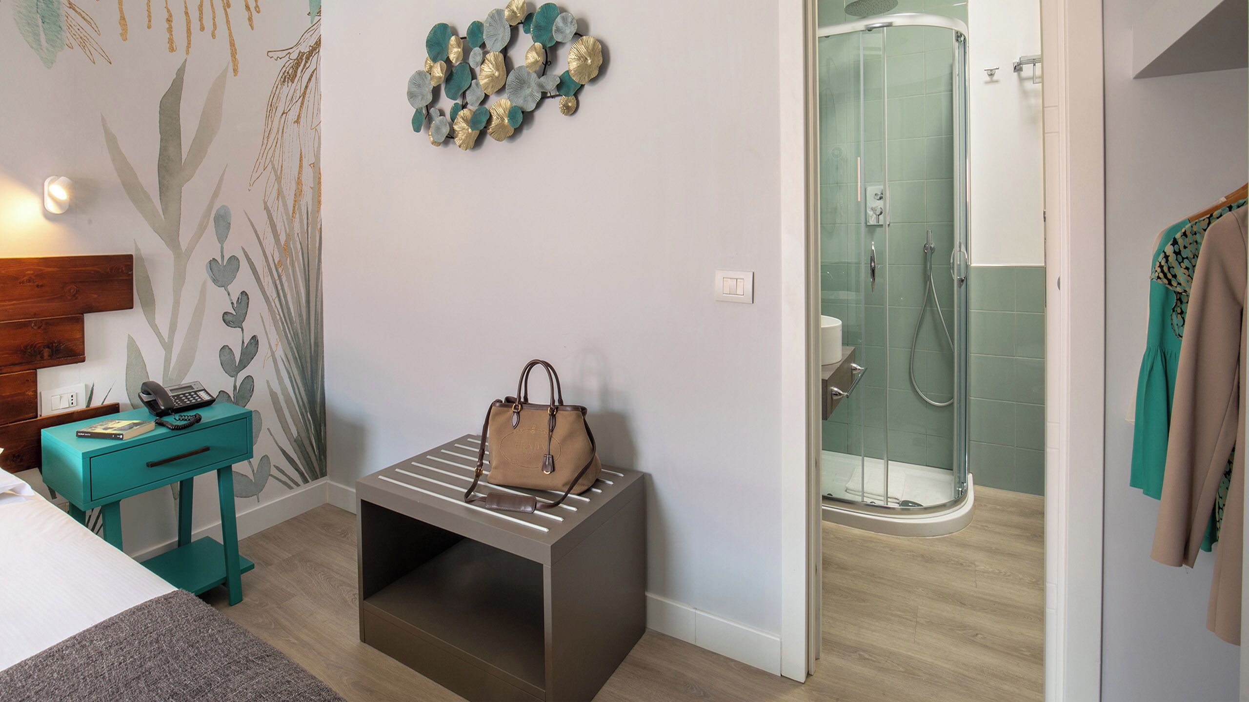 A hotel room with a bed, a teal nightstand with a phone and book, a brown handbag on a bench, a shower area, and a coat rack with a coat.