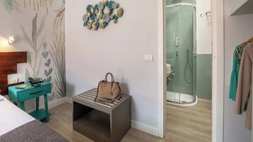 A hotel room with a bed, a teal nightstand with a phone and book, a brown handbag on a bench, a shower area, and a coat rack with a coat.