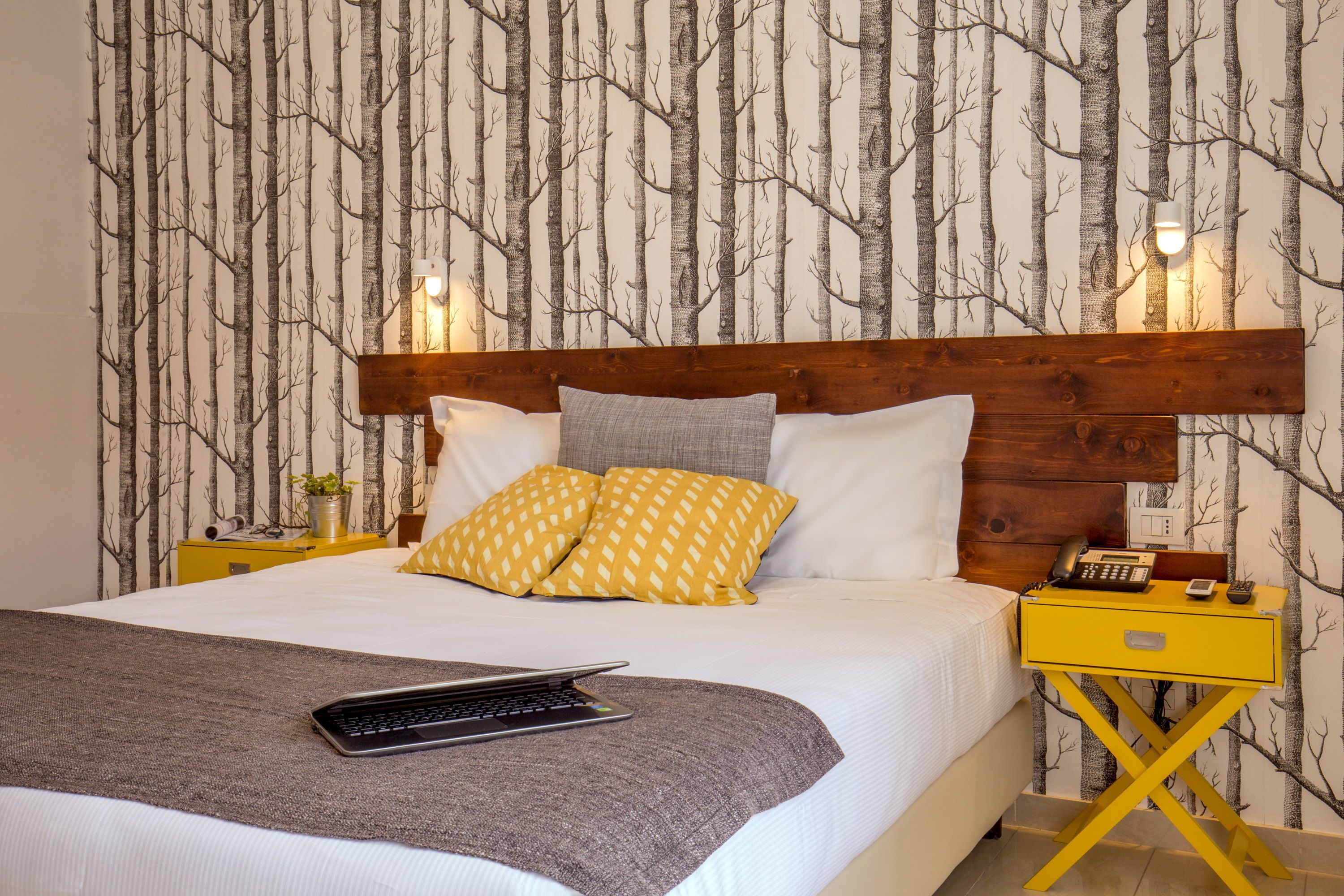 Un lit bien fait, avec une tête de lit en bois, deux coussins à motifs jaunes, un coussin gris et un ordinateur portable posé sur un plaid gris.