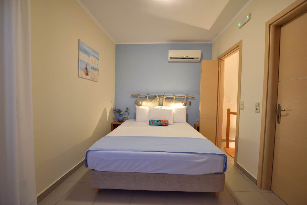 A hotel room with a bed, a nightstand, an air conditioning unit, and a door.