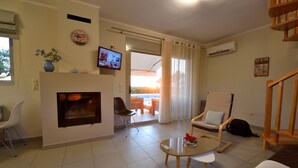 A living room with a fireplace, a television, a dining area with a table and chairs, and a view through a door. - Edem Suites (Trifylia)