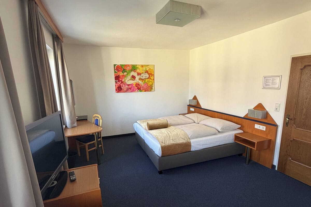 Habitación de hotel con cama, escritorio con televisor, una silla, un cuadro en la pared y una puerta.