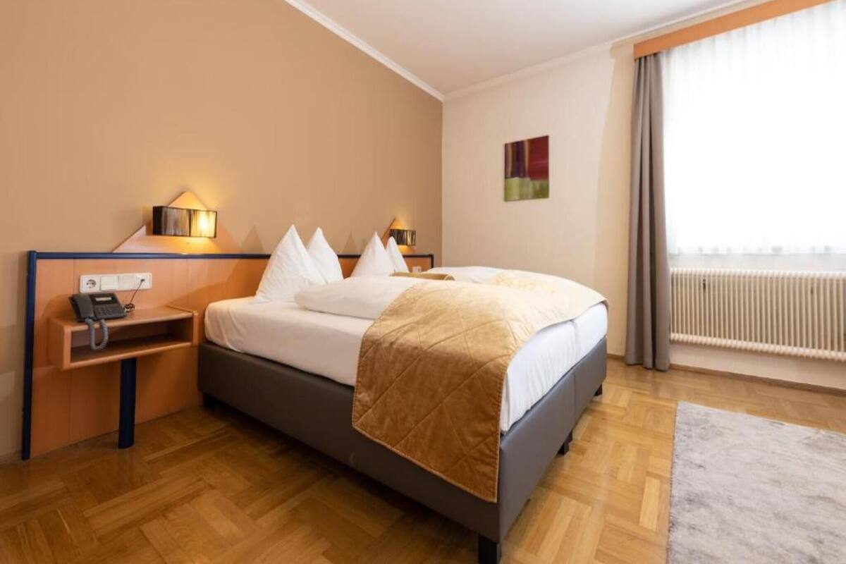 Una habitación de hotel con cama, mesitas de noche, teléfono, lámpara, ventana con cortinas y radiador.