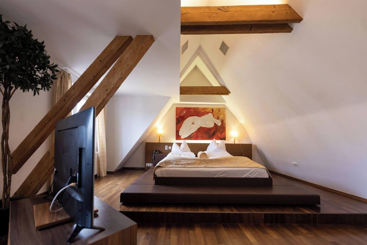 Un dormitorio moderno con techo inclinado, cama con ropa de cama blanca, un cuadro en la pared y un televisor sobre un soporte de madera.
