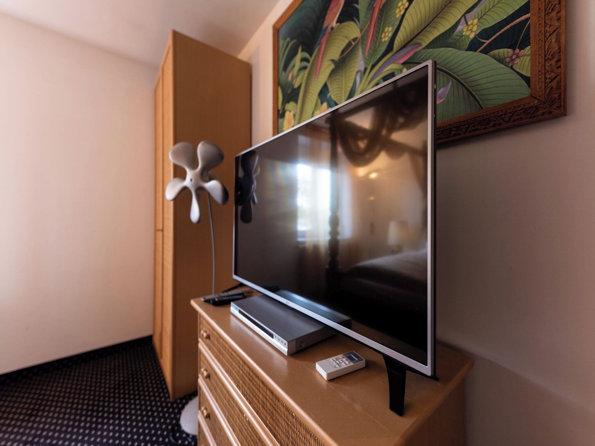 A modern hotel room with a flat-screen TV on a wooden dresser, a remote control, and a decorative fan.