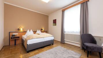Ein Hotelzimmer mit Bett, Nachttischen, einem Sessel, einem Fenster mit Vorhängen, einer Heizung und einer Leuchte.