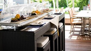 Ein Buffet mit transparenten Abdeckungen über den Speisen, Sitzgelegenheiten im Freien mit Tischen und Stühlen und ein Blick auf Grün im Hintergrund.