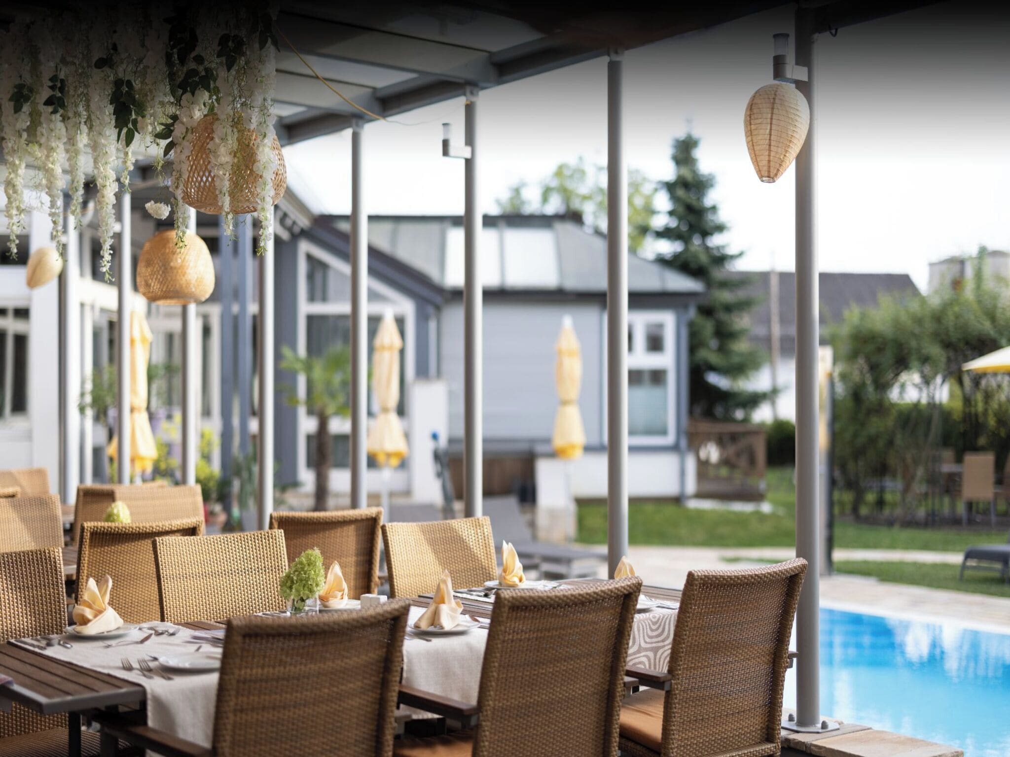 An outdoor dining area with wicker furniture, hanging decorations, and a view of a swimming pool and buildings.