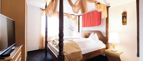A four-poster bed with a canopy, a television, a desk with drawers, a chair, and a lamp.