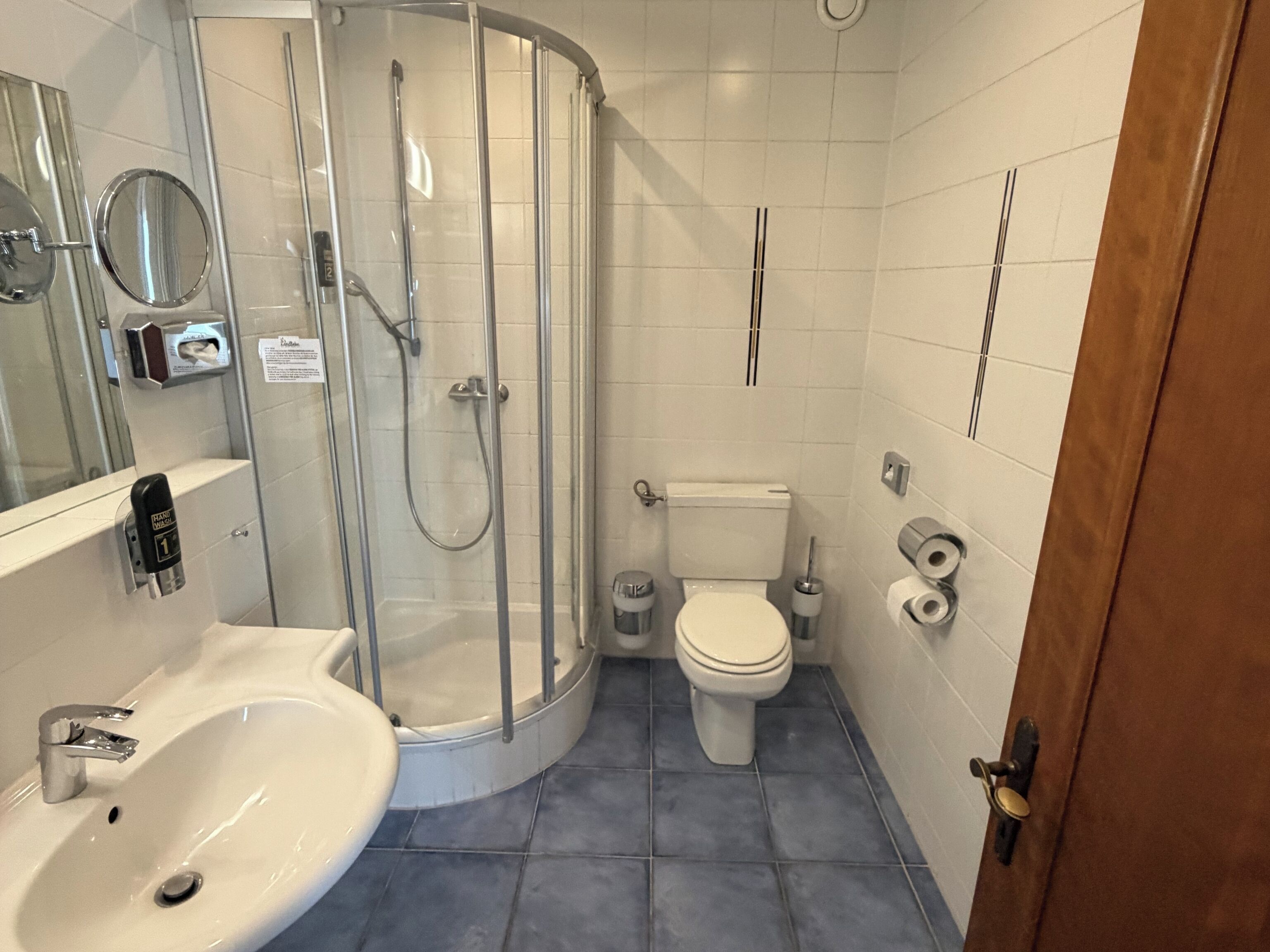 A bathroom with a glass-enclosed shower, a white sink, a toilet, and a door with a brass handle.