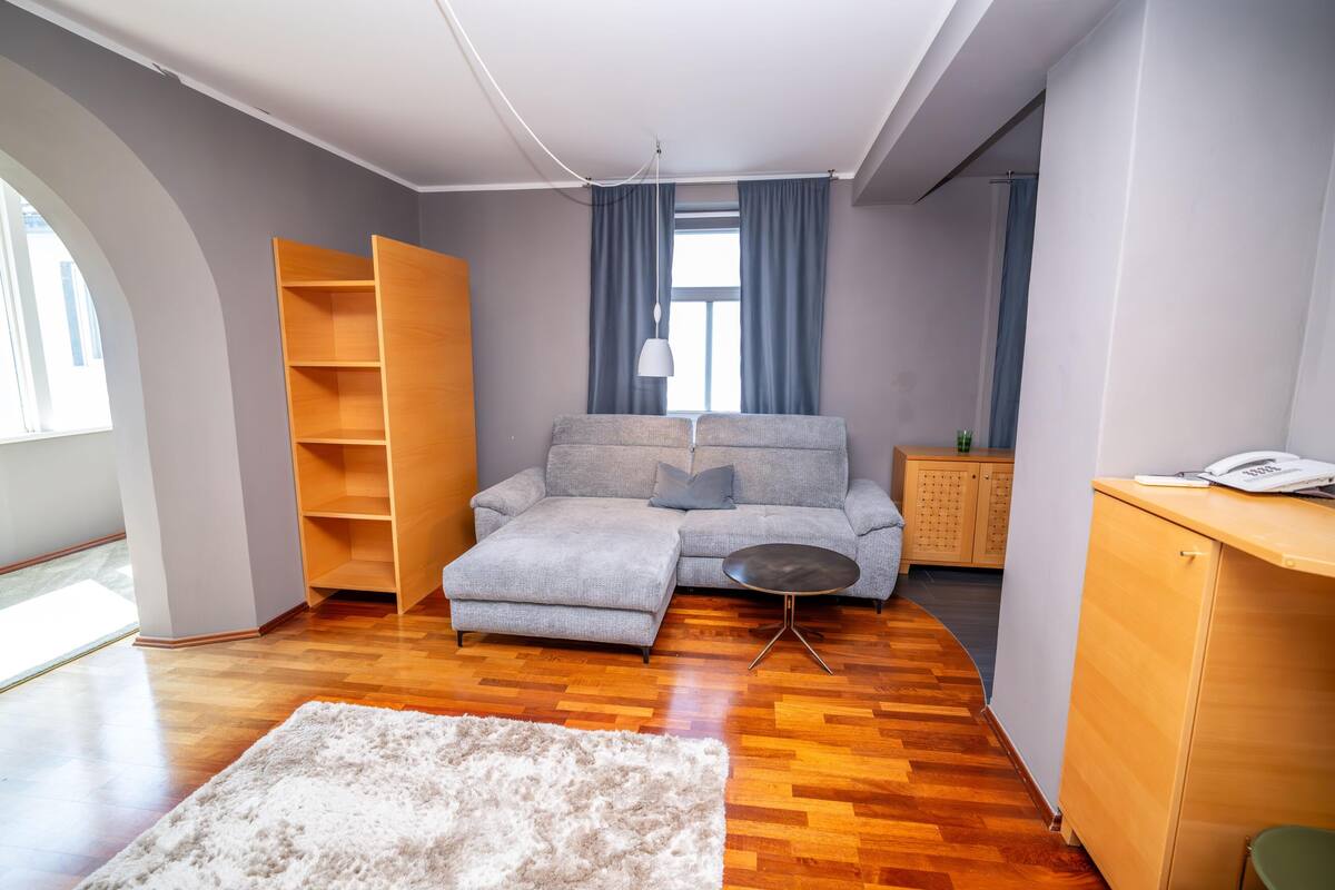 Una sala moderna con un sofá seccional gris, una mesita redonda, un mueble de madera y un ventanal con cortinas.