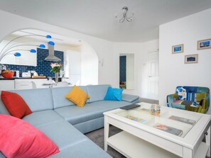 Een moderne woonkamer met een blauwe bank, een witte salontafel en een kleurrijke fauteuil. Op de achtergrond is een keuken met blauwe tegels en witte kastjes te zien.