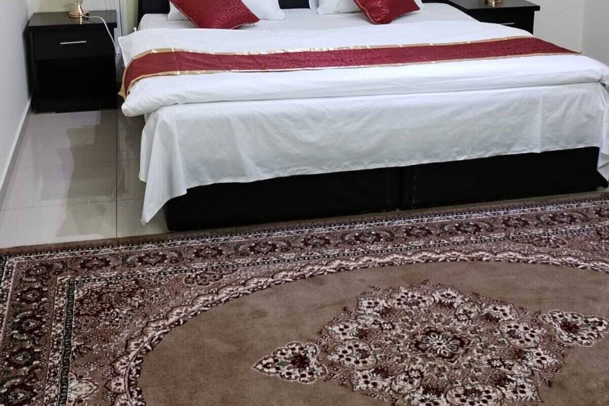 Cama bem arrumada com colcha branca e vermelha, duas almofadas vermelhas e cabeceira escura com design decorativo. Há duas mesas de cabeceira com luminárias, um tapete estampado no chão e uma janela com cortinas.