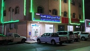 A multi-story building with green neon lights outlining its structure. There are several parked vehicles in front, and a sign indicating the building is a serviced apartment complex.