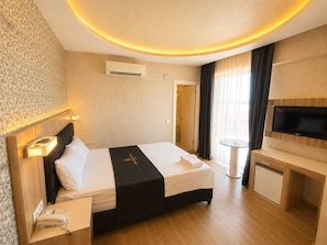 Habitación de hotel con cama, mesita de noche, lámpara, aire acondicionado, televisor y un escritorio pequeño con una silla.