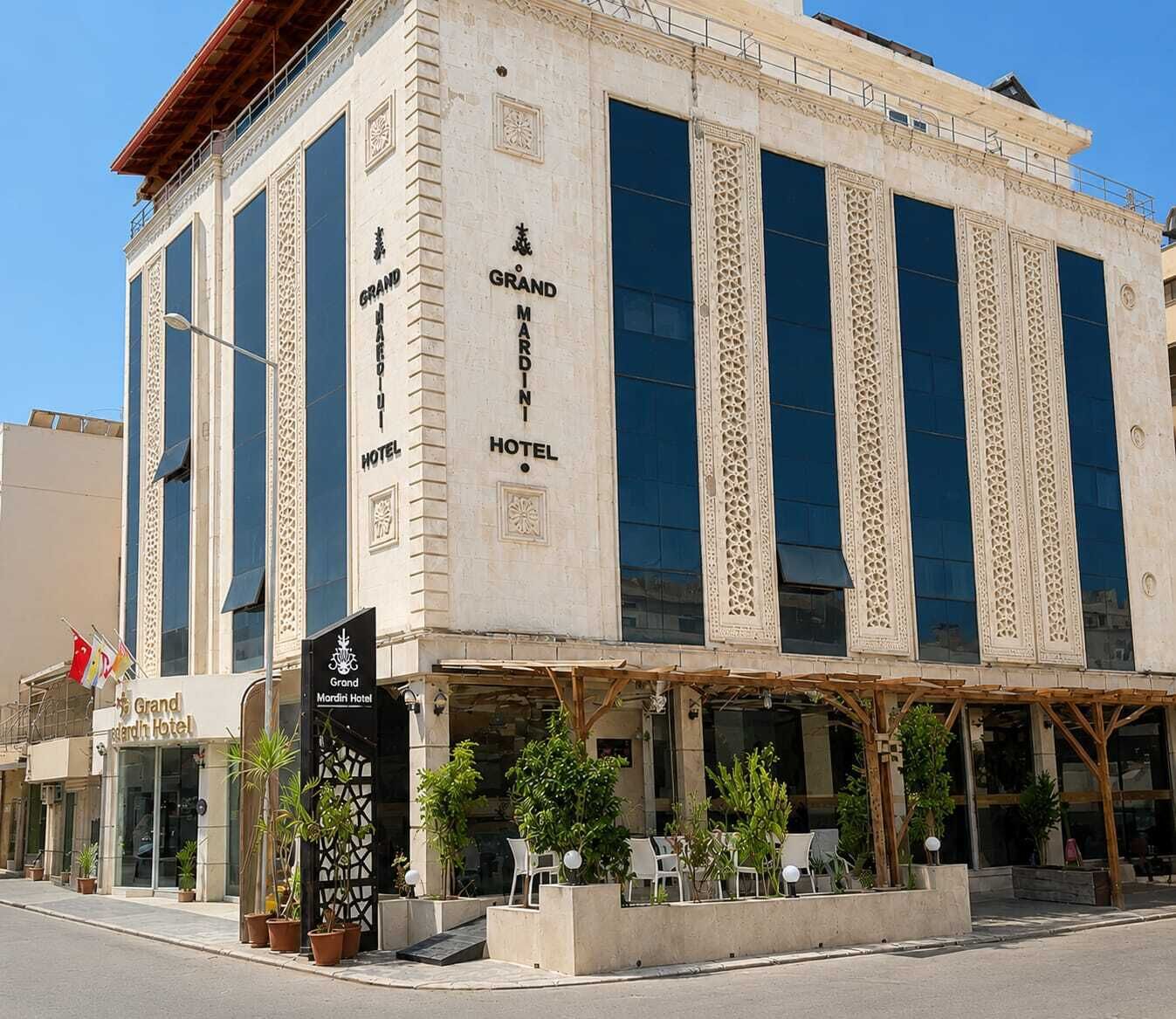 Un hôtel à la façade classique, doté de grandes fenêtres et d’un espace extérieur aménagé avec des plantes, où l’on peut s’asseoir à l’abri.