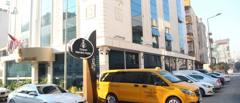A Grand Mariani Hotel with a modern facade, a yellow van parked in front, and several cars parked along the street.