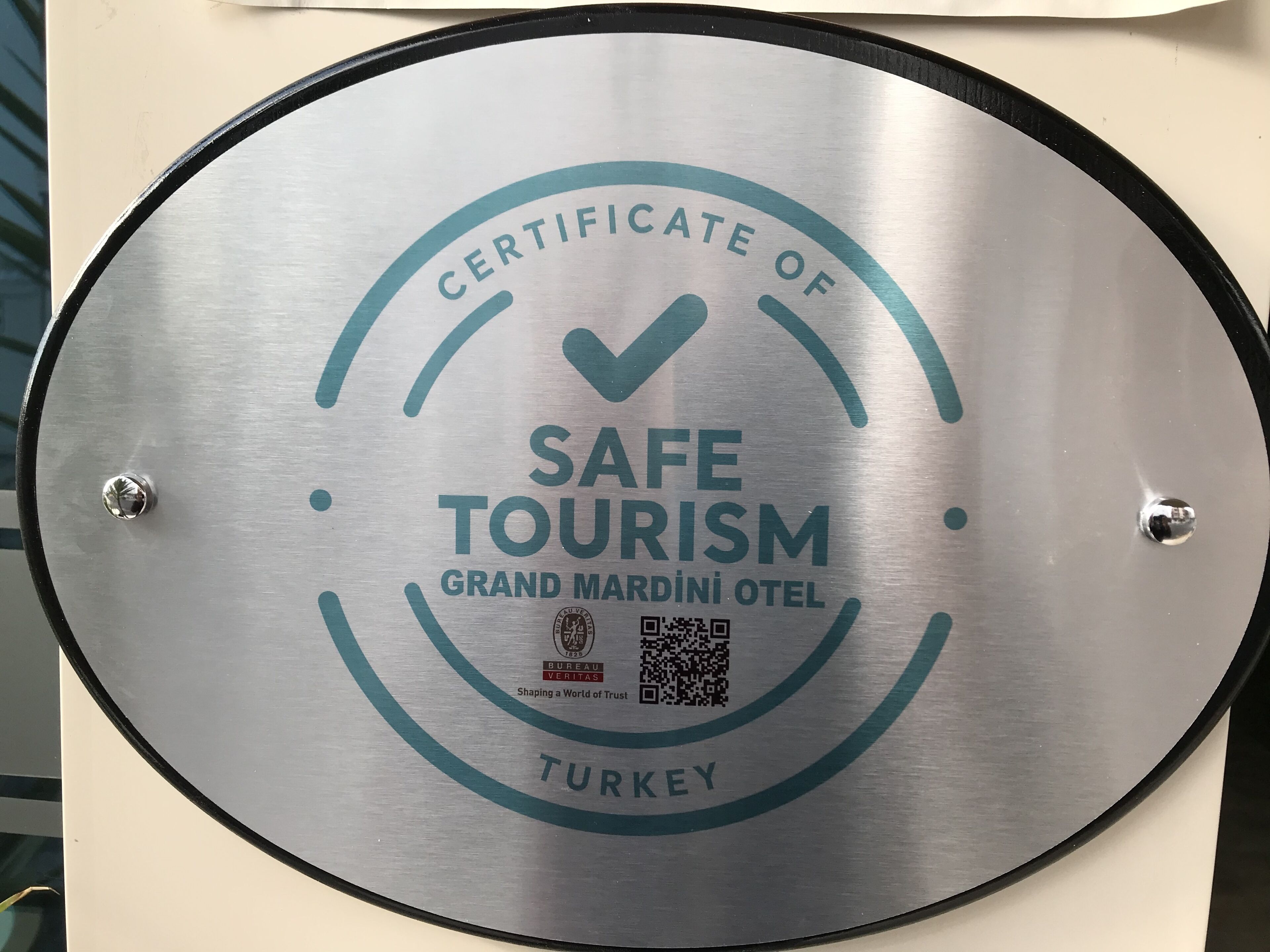 A circular sign with the text "CERTIFICATE OF SAFE TOURISM GRAND MARDINI OTEL TURKEY" and a QR code.