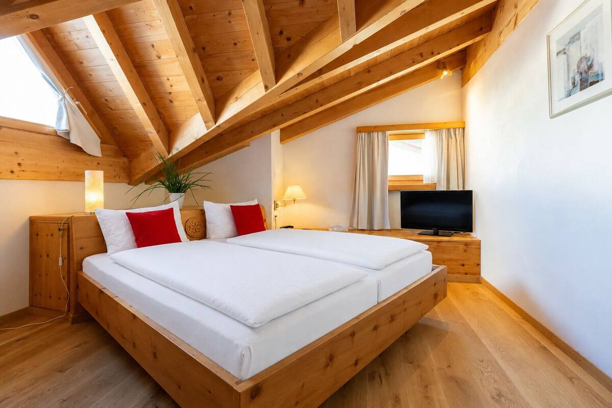 Une chambre avec un plafond en bois, un lit avec une literie blanche et rouge, une télévision et une fenêtre avec des rideaux.