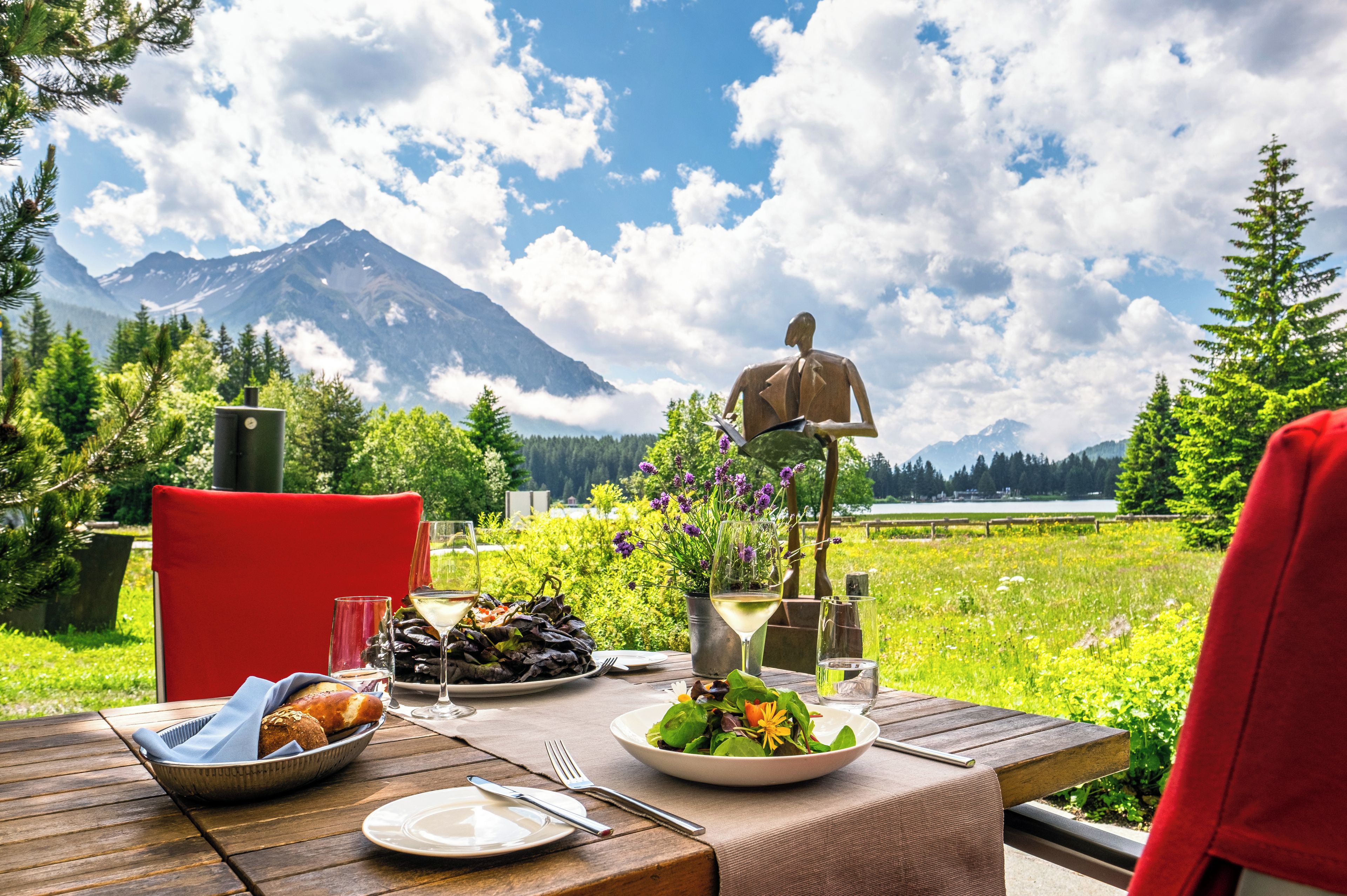 A dining table set outdoors with a mountain view, a statue, and a lake in the background.
