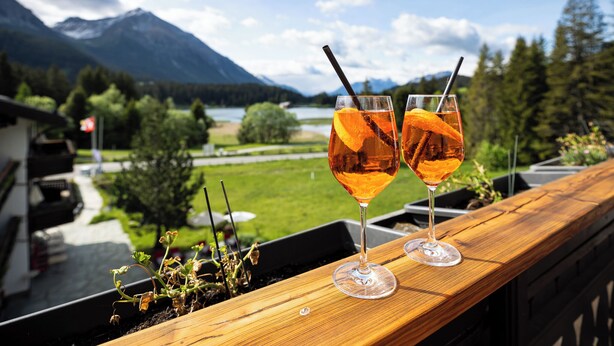 Two glasses of amber liquid with orange slices and straws on a wooden ledge with a scenic mountain and lake view.