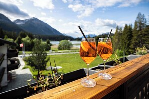 Zwei Gläser bernsteinfarbige Flüssigkeit mit Orangenscheiben und Strohhalmen auf einer Holzablage, mit Blick auf Berge und einen See.