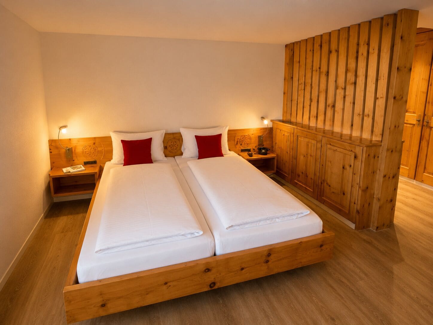 Cama doble con sábanas blancas, dos almohadas rojas, cabecera de madera, mesitas de noche con lámparas y un armario de madera.
