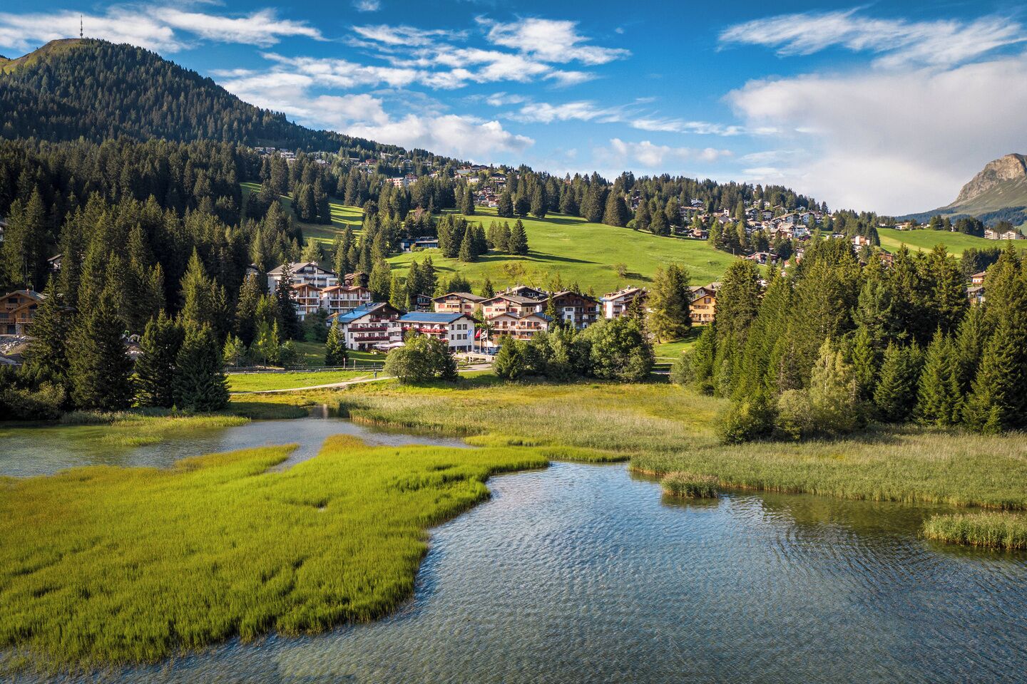 Un pueblo a orillas de un lago, con edificios rodeados de vegetación y una montaña al fondo.