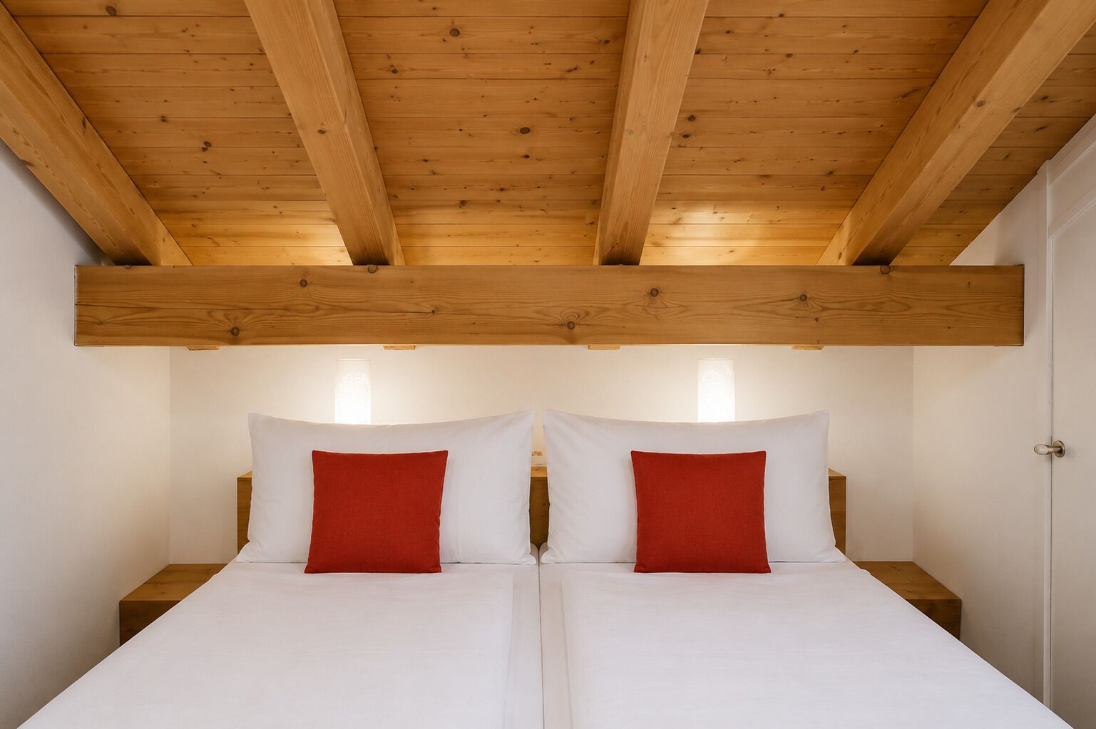 A double bed with white bedding and red pillows, wooden beams on the ceiling, and bedside tables.