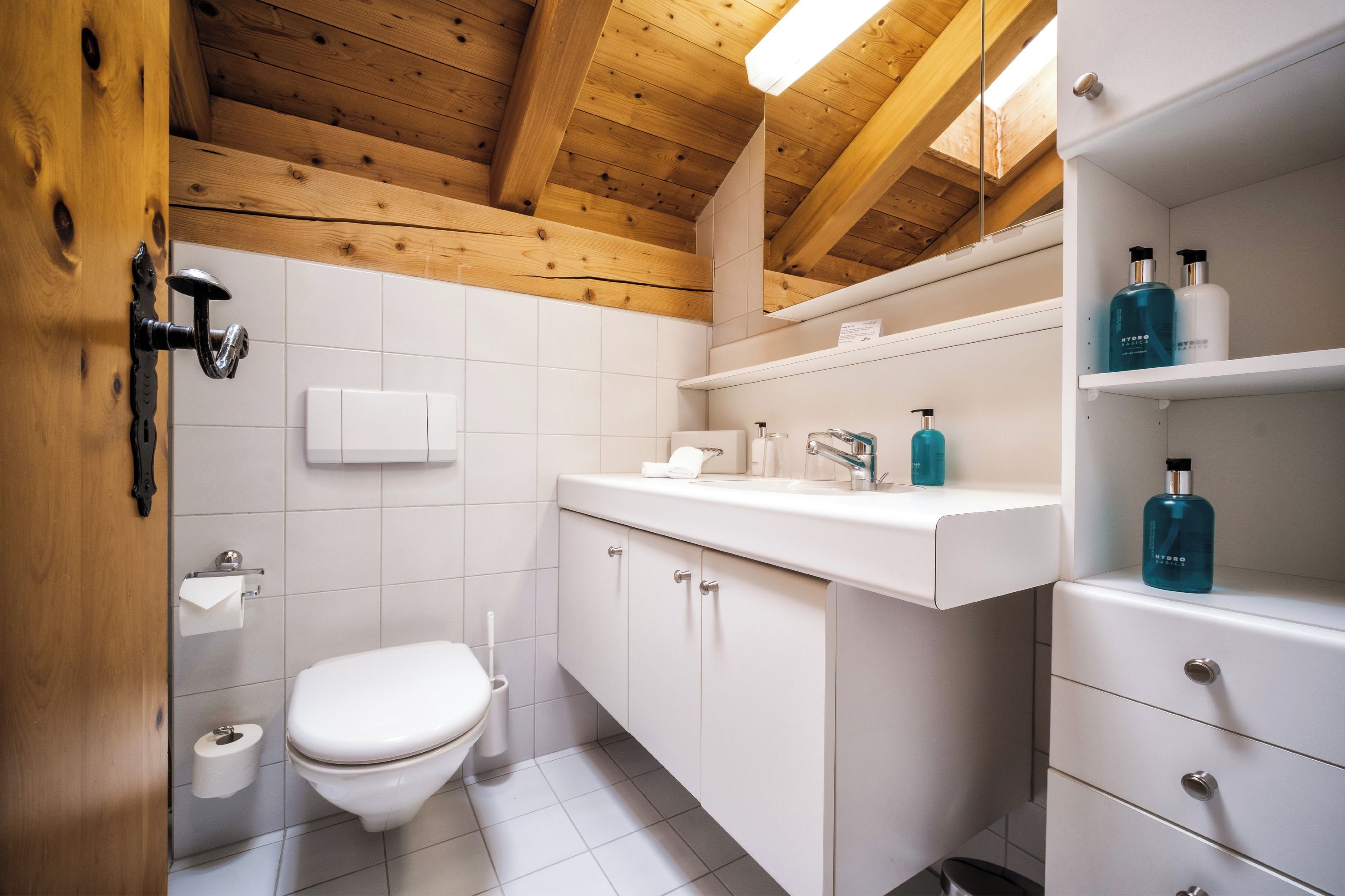Ein modernes Badezimmer mit Holzboden, weißen Fliesen, einer Toilette, einem Waschbecken mit Spiegel und Pflegeprodukten auf einem Regal.