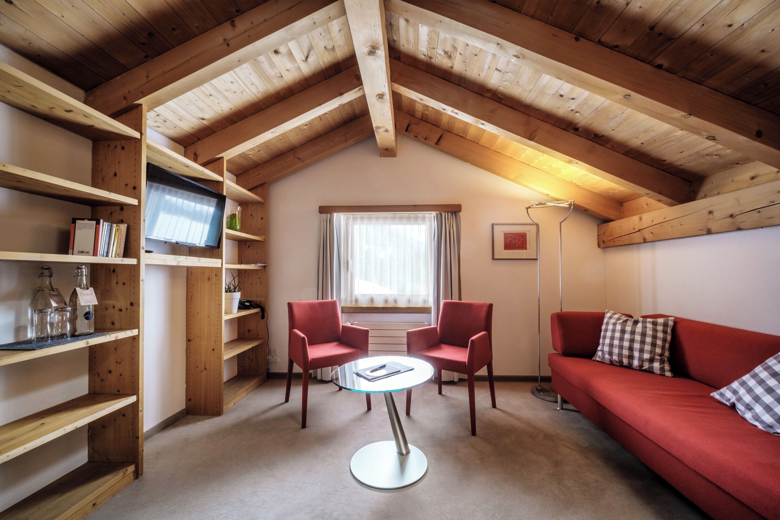 A cozy room with a wooden ceiling, a red sofa, two red chairs, a glass table, and a TV on a shelf.