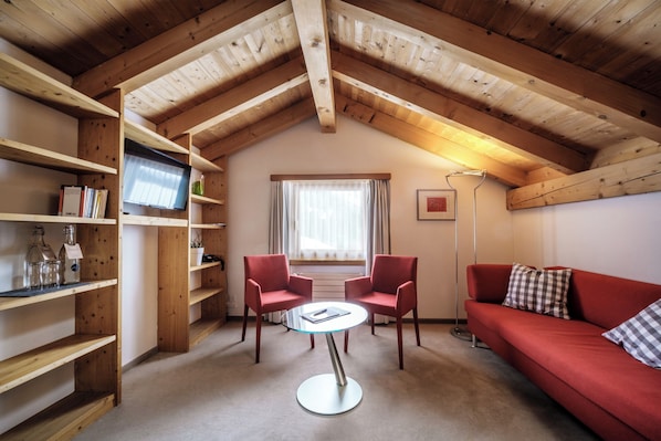Una habitación acogedora con techo de madera, un sofá rojo, dos sillas rojas, una mesa de vidrio y un televisor en un estante.
