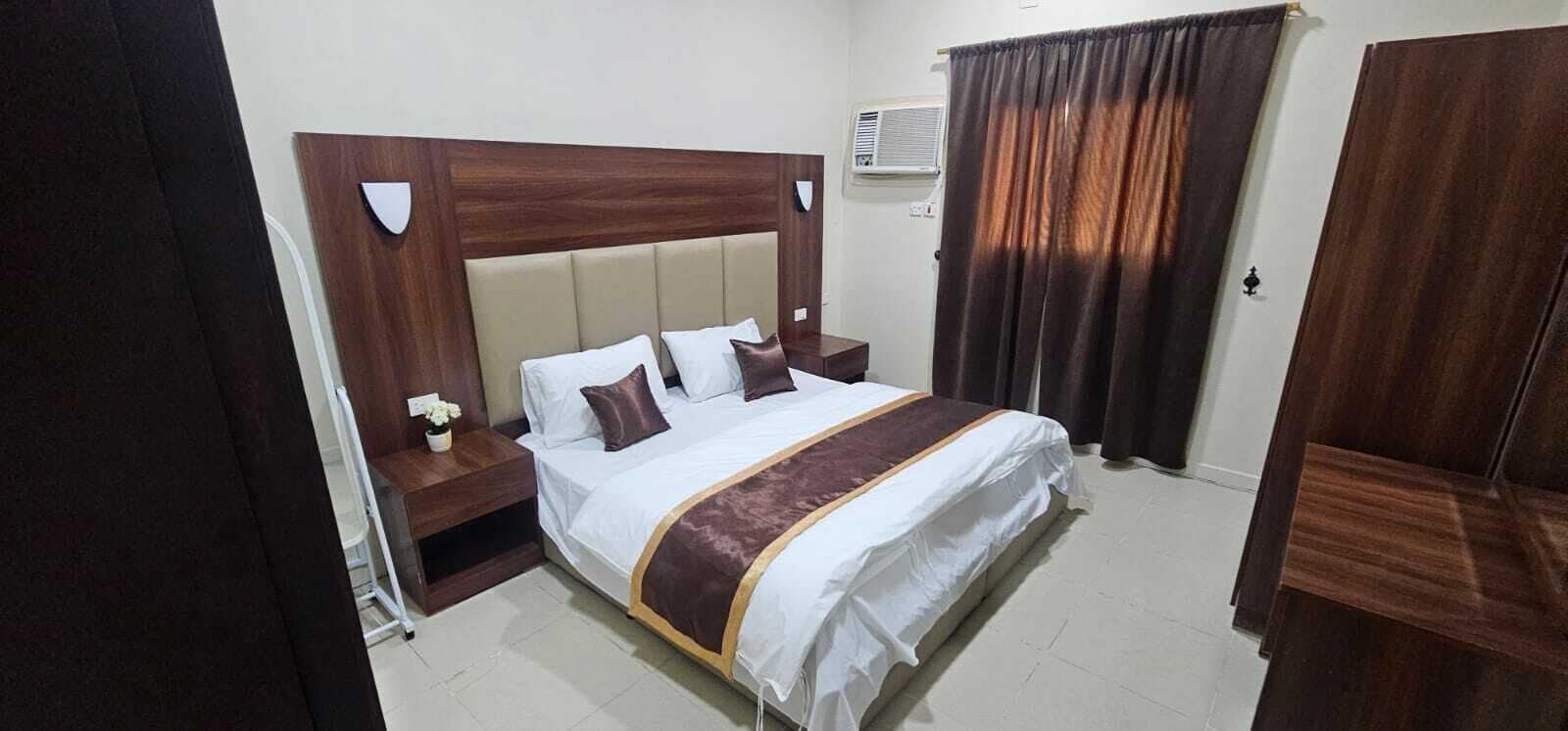 A hotel room with a large bed, a wooden headboard, a nightstand with a lamp, and a window with curtains.