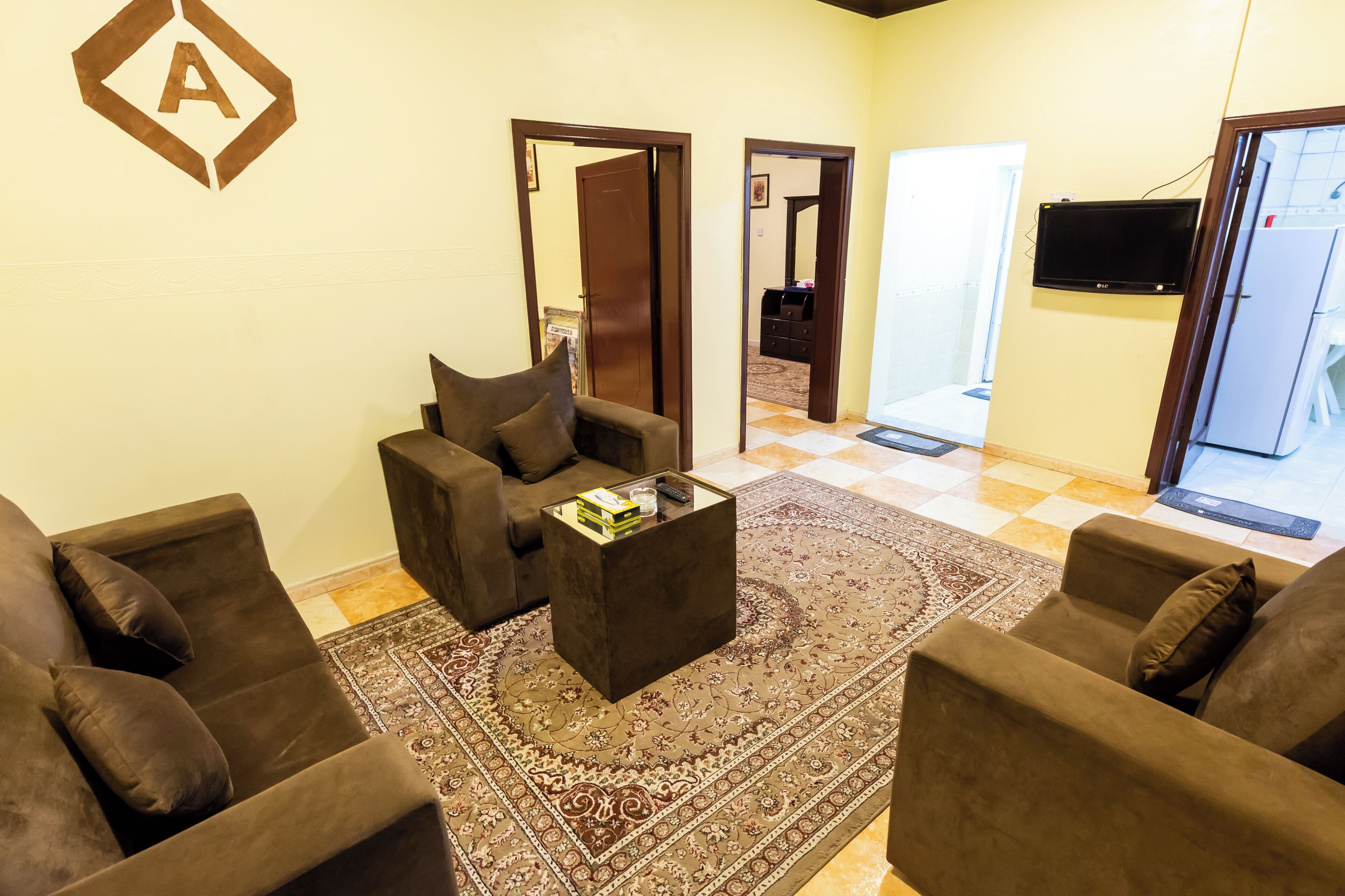 Un salon avec un ensemble de canapés marron, une table basse et un tapis à motifs. Une porte donne accès à une autre pièce et un téléviseur est fixé au mur.