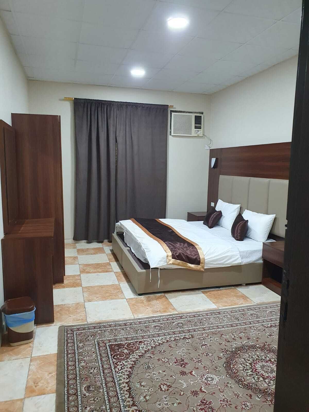 Un dormitorio con cama, mesita de noche, armario de madera, cortina y alfombra estampada.