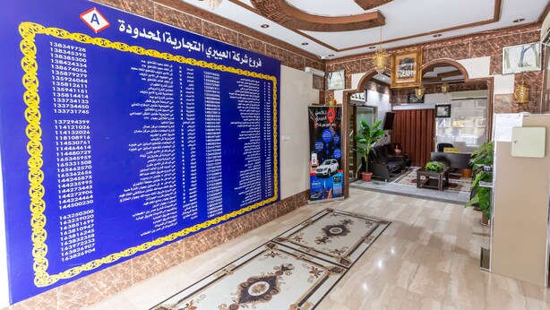 A hotel lobby with a large blue wall displaying room numbers and names, a chandelier, and a seating area with a coffee table and chairs.