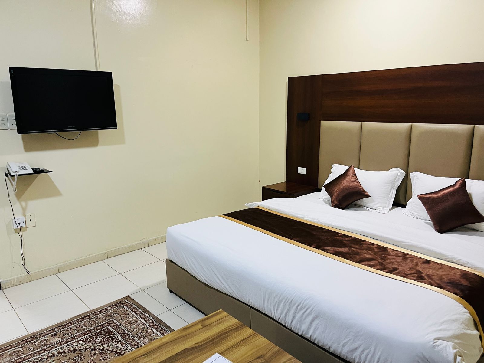 A hotel room with a bed, a television mounted on the wall, a bedside table, and a small rug.