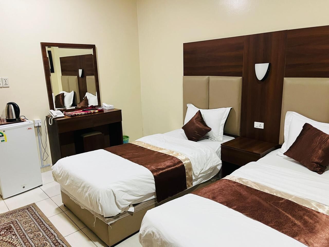 A hotel room with two beds, a desk with a telephone, a mirror, and a refrigerator.
