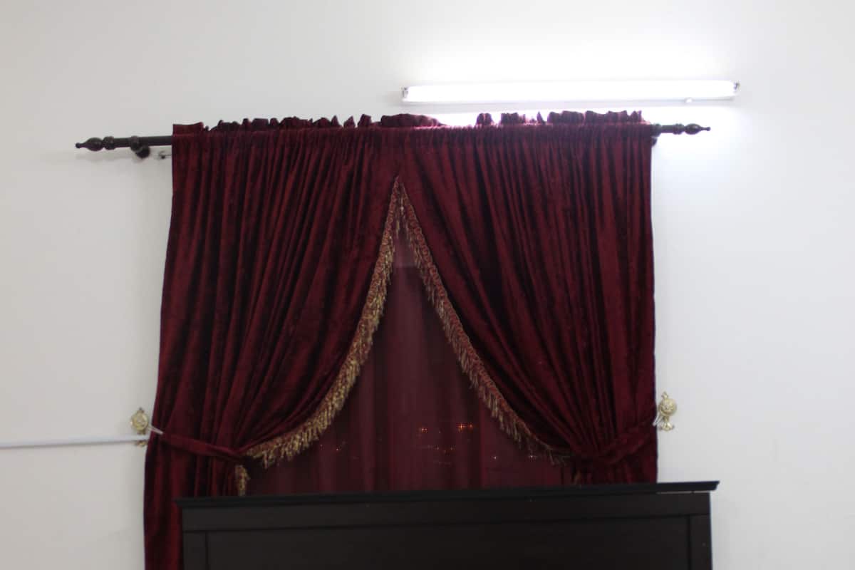 Un lit bien fait, avec du linge de lit blanc et deux coussins décoratifs, une tête de lit en bois foncé et un rideau rouge orné d’une bordure dorée.