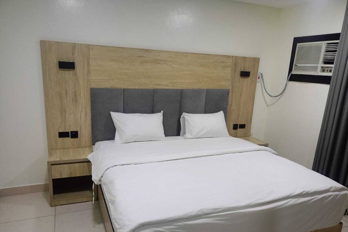 Habitación de hotel con cabecera de madera, dos almohadas, una colcha blanca y una mesita de noche pequeña.