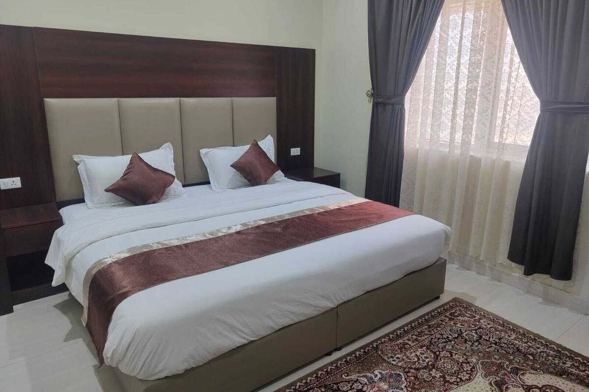 Habitación de hotel con una cama grande, cabecera de madera, mesita de noche y ventana con cortinas.