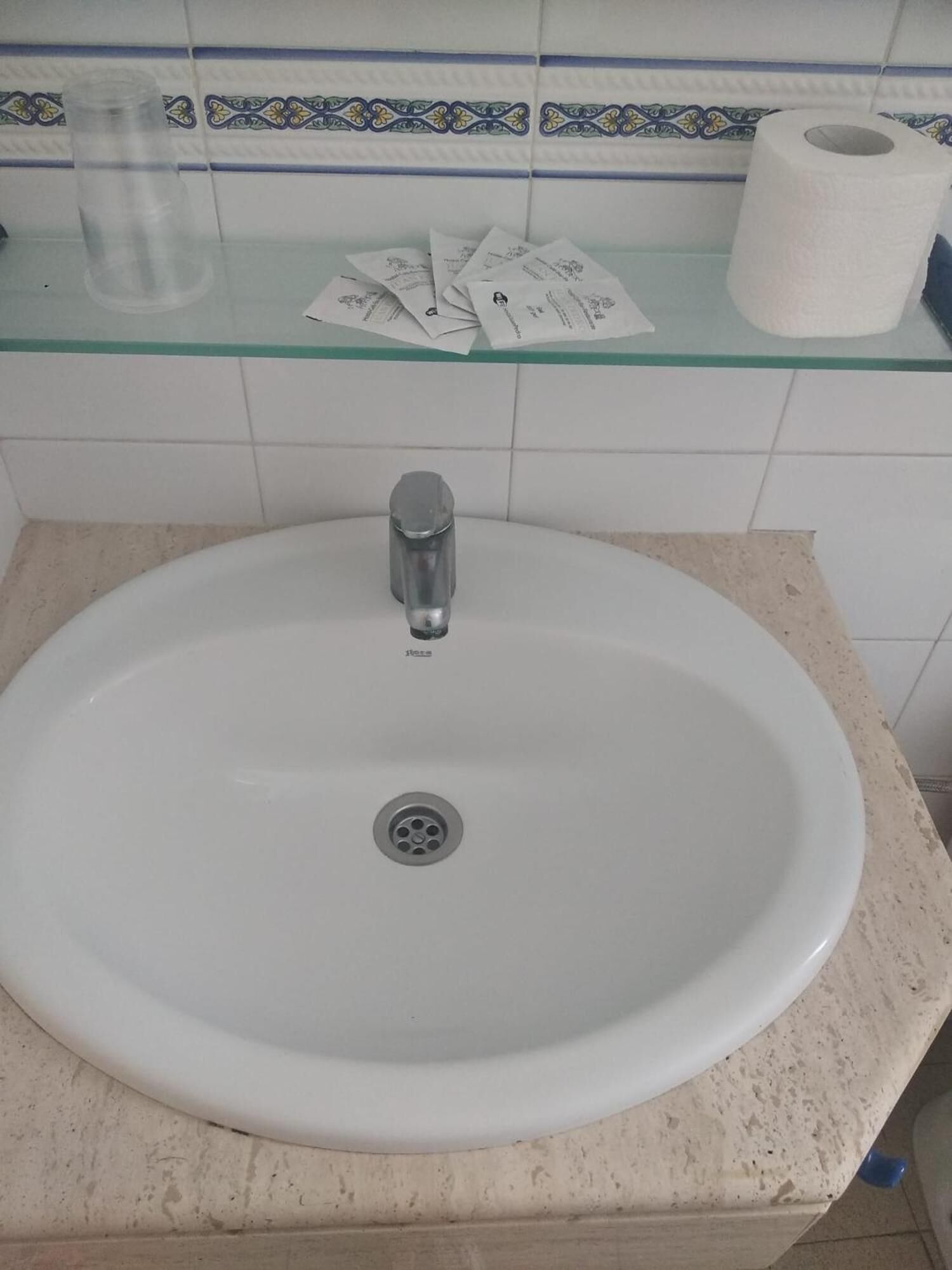 A bathroom sink with a faucet, a glass container, and a roll of toilet paper on a shelf.
