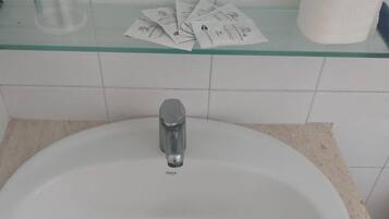 A bathroom sink with a faucet, a glass container, and a roll of toilet paper on a shelf.