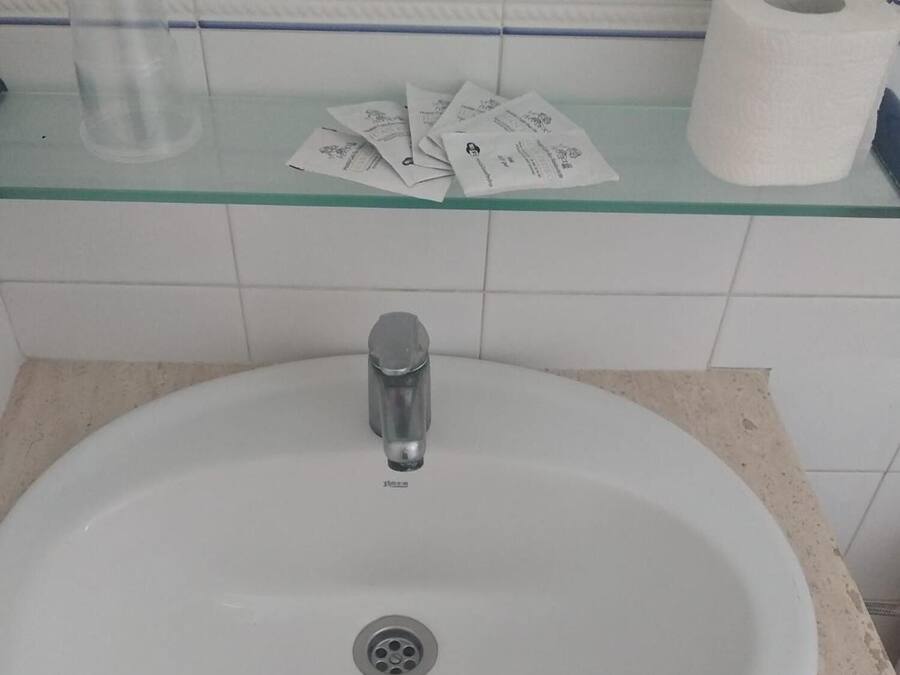 A bathroom sink with a faucet, a glass container, and a roll of toilet paper on a shelf.
