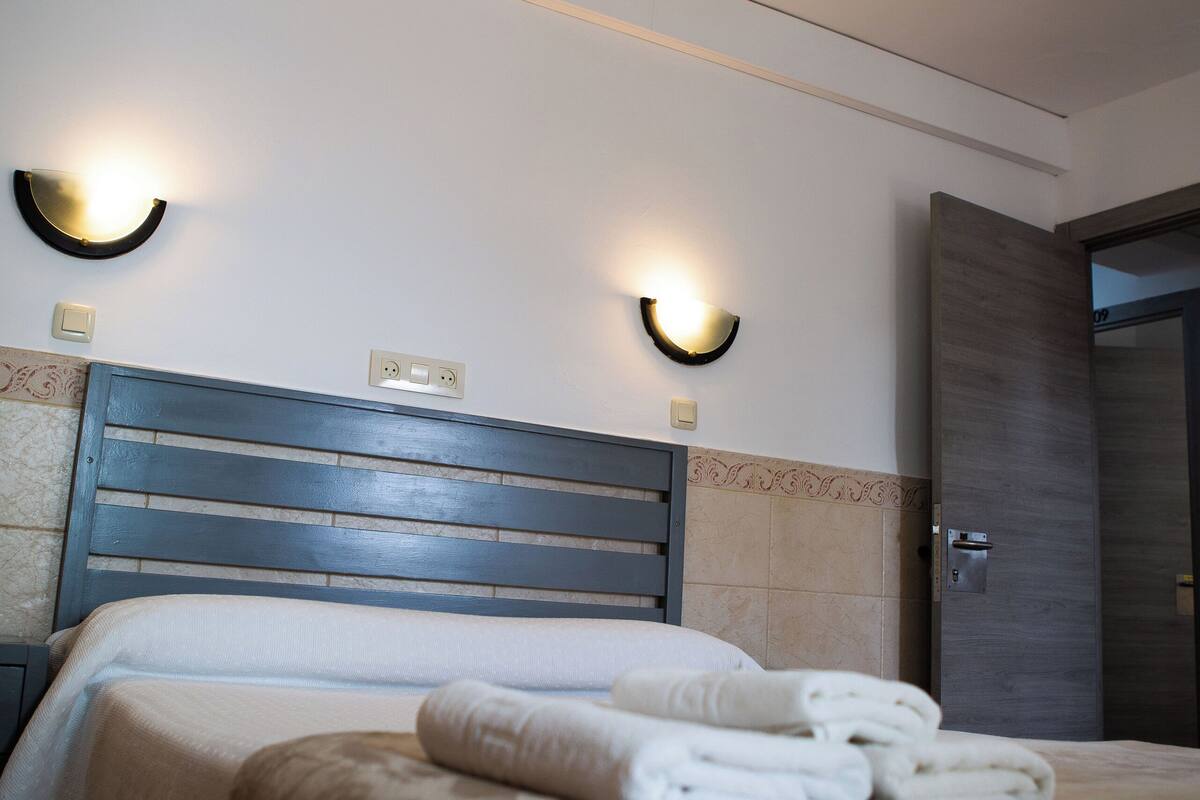 A hotel room with a bed, two wall-mounted lights, a door, and a wall socket.