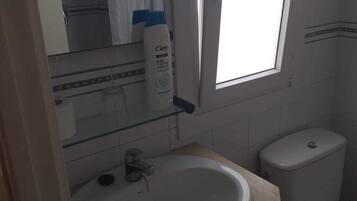 Un baño con lavamanos blanco, inodoro y estante de vidrio con artículos de aseo.