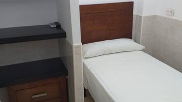 Una habitación pequeña y limpia con una cama individual, un aparador de madera con cajones y una estantería encima.